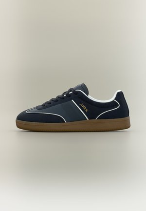 Pantof sport negru, model cu talpă joasă, cu margine albă, talpă maro și inscripție aurie „ARKK” pe lateral, pe fundal neutru.