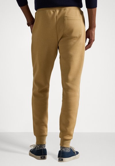 Beige cotton sweatpants with elastic cuffs, featuring a back pocket and a smooth texture, worn with navy shoes and patterned socks.