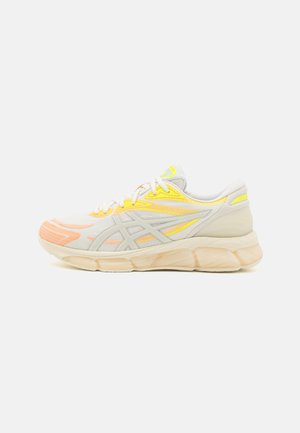 Chaussure de sport beige clair avec dessus en mesh, accents jaunes et pêche, lacets blancs, semelle épaisse et rembourrée, et bandes latérales de la marque, vue de profil.