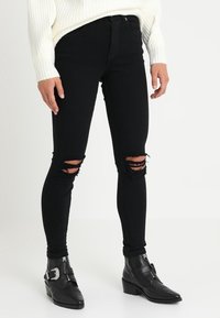 Svarta skinny jeans med slitningar vid knäna, åtföljda av svarta ankelskor med spänne och spetsig tå. Vit stickad tröja synlig.