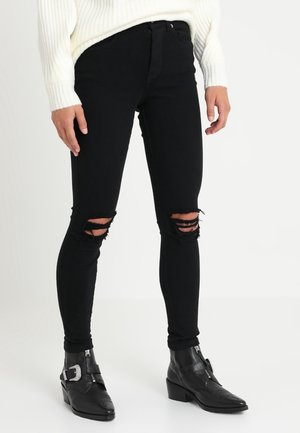 Jeans negros ajustados con desgastes en las rodillas, combinados con botines negros de tobillo con hebilla y punta afilada. Suéter blanco de punto visible.