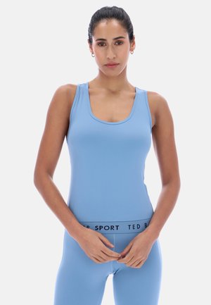 Kobieta o ciemnych włosach, ubrana w niebieski sportowy top bez rękawów oraz pasujące legginsy z napisem "TED BAKER SPORT" na talii, stojąca na jednolitym tle.