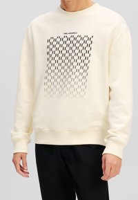 Felpa color crema con maniche lunghe, polsini e orlo a costine. Presenta un motivo geometrico nero e il testo "KARL LAGERFELD" sopra il disegno.