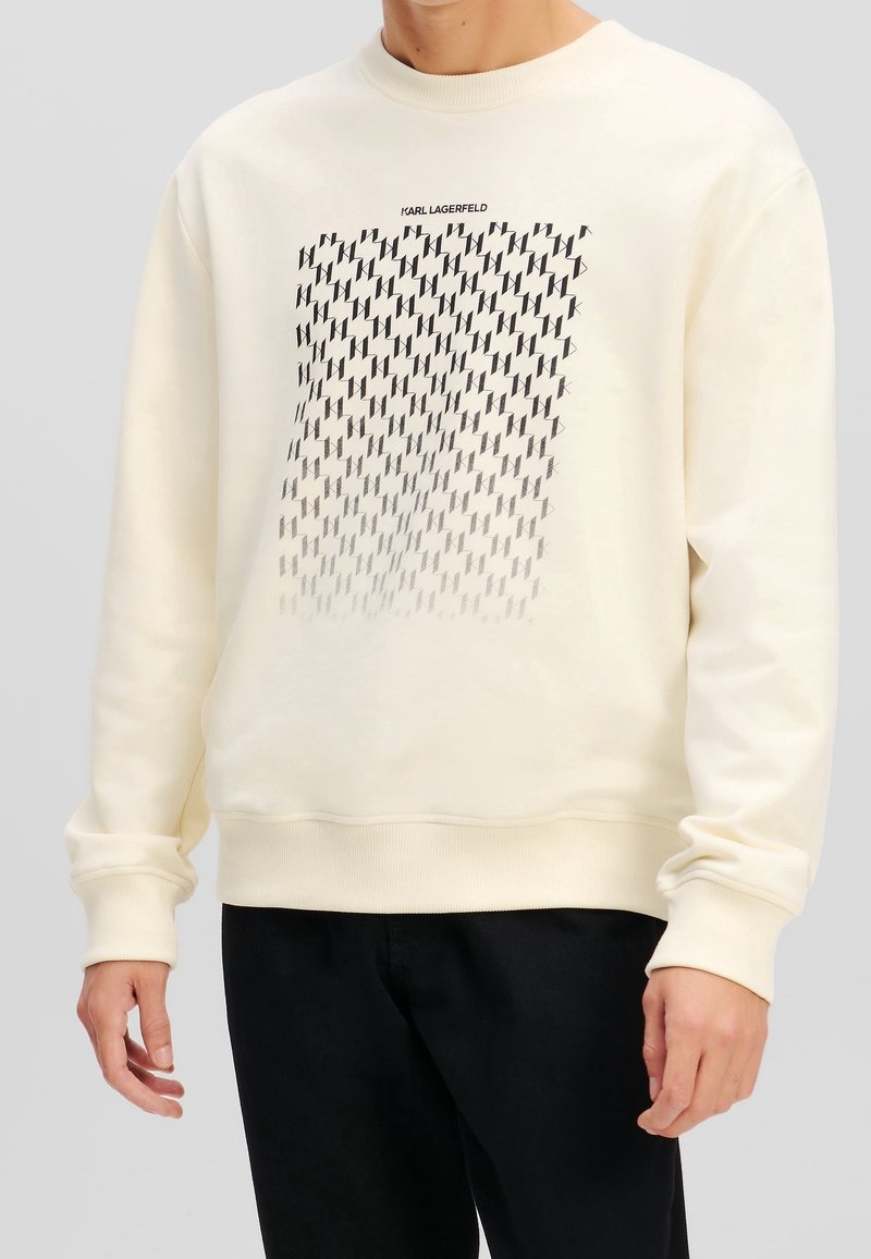 Felpa color crema con maniche lunghe, polsini e orlo a costine. Presenta un motivo geometrico nero e il testo "KARL LAGERFELD" sopra il disegno.