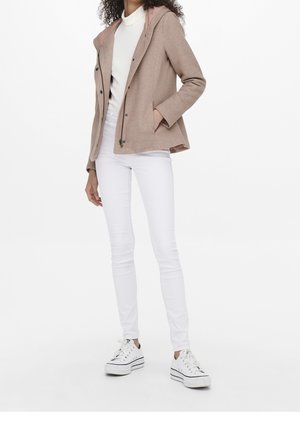 Femme portant une veste à capuche beige, un col roulé blanc, un pantalon blanc moulant et des baskets compensées blanches, debout avec une main dans la poche.