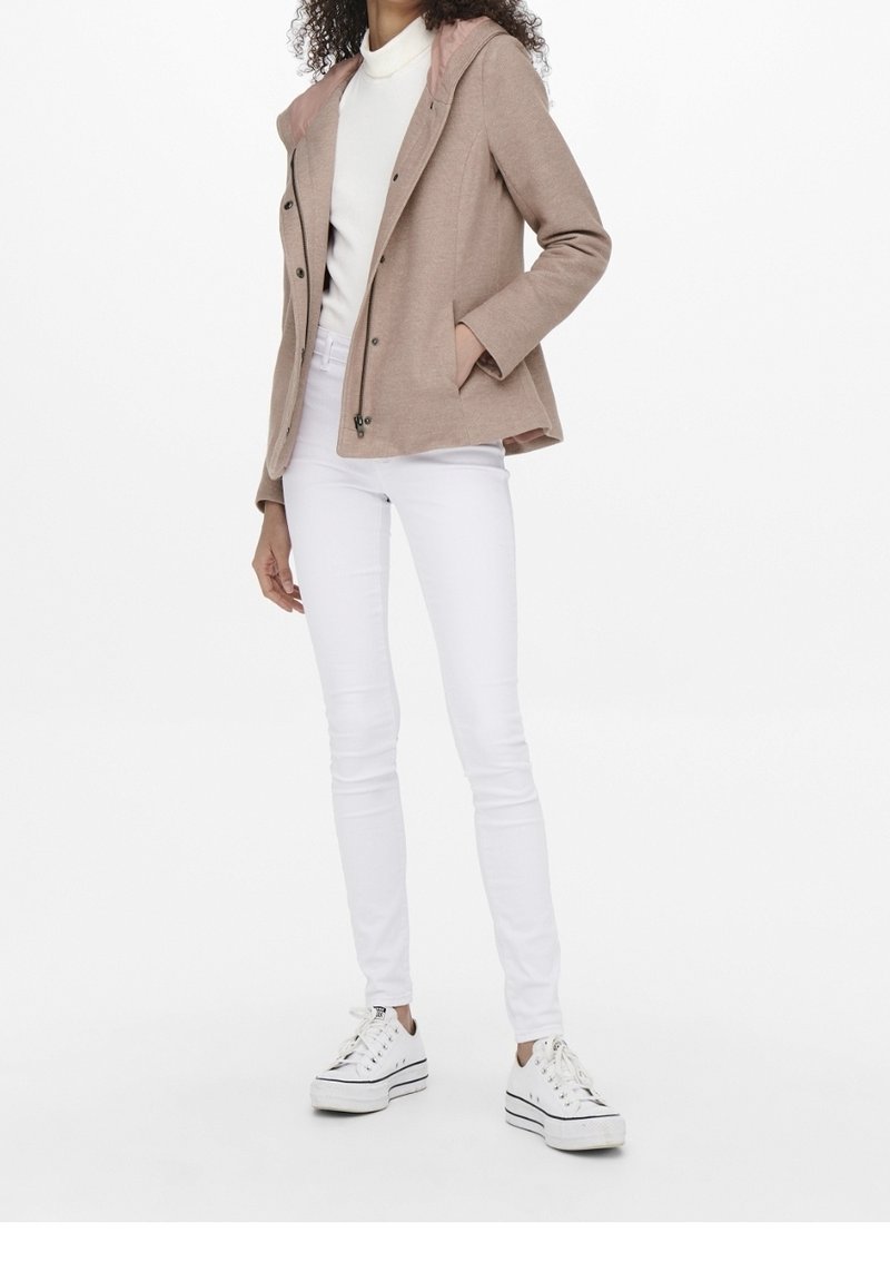 Femme portant une veste à capuche beige, un col roulé blanc, un pantalon blanc moulant et des baskets compensées blanches, debout avec une main dans la poche.