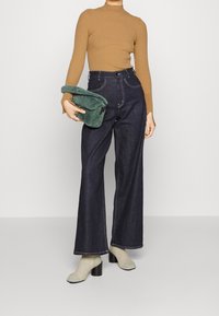 Geribbelde bruine lange mouw top, donkerblauwe hoge taille wijde jeans, groene pluche tas en beige gestructureerde sokken met zwarte blokhakken.