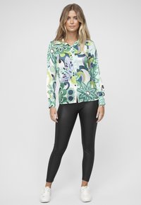 Košile s dlouhým rukávem na knoflíky se zeleno-fialovým květinovým vzorem, vyrobená z lesklé látky, kombinovaná s černými legínami a bílými teniskami.