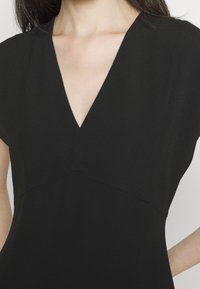 Svart klänning med V-ringning och korta ärmar. Slät, texturerad tyg med figurnära liv och en flödande nederdel.