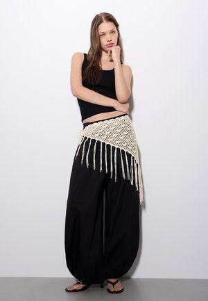 Junge Frau mit langen Haaren in schwarzem Tanktop, weiten schwarzen Hosen und cremefarbenem gehäkeltem Fransen-Gürtel, steht vor einer weißen Wand mit der Hand am Kinn.