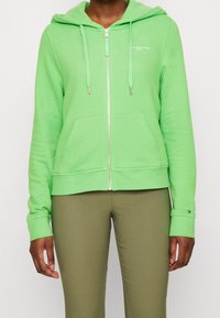 Sudadera verde con cremallera, hecha de tejido suave, que cuenta con un bolsillo frontal, cordones y herrajes en tono dorado. Llevada con pantalones verde oliva.