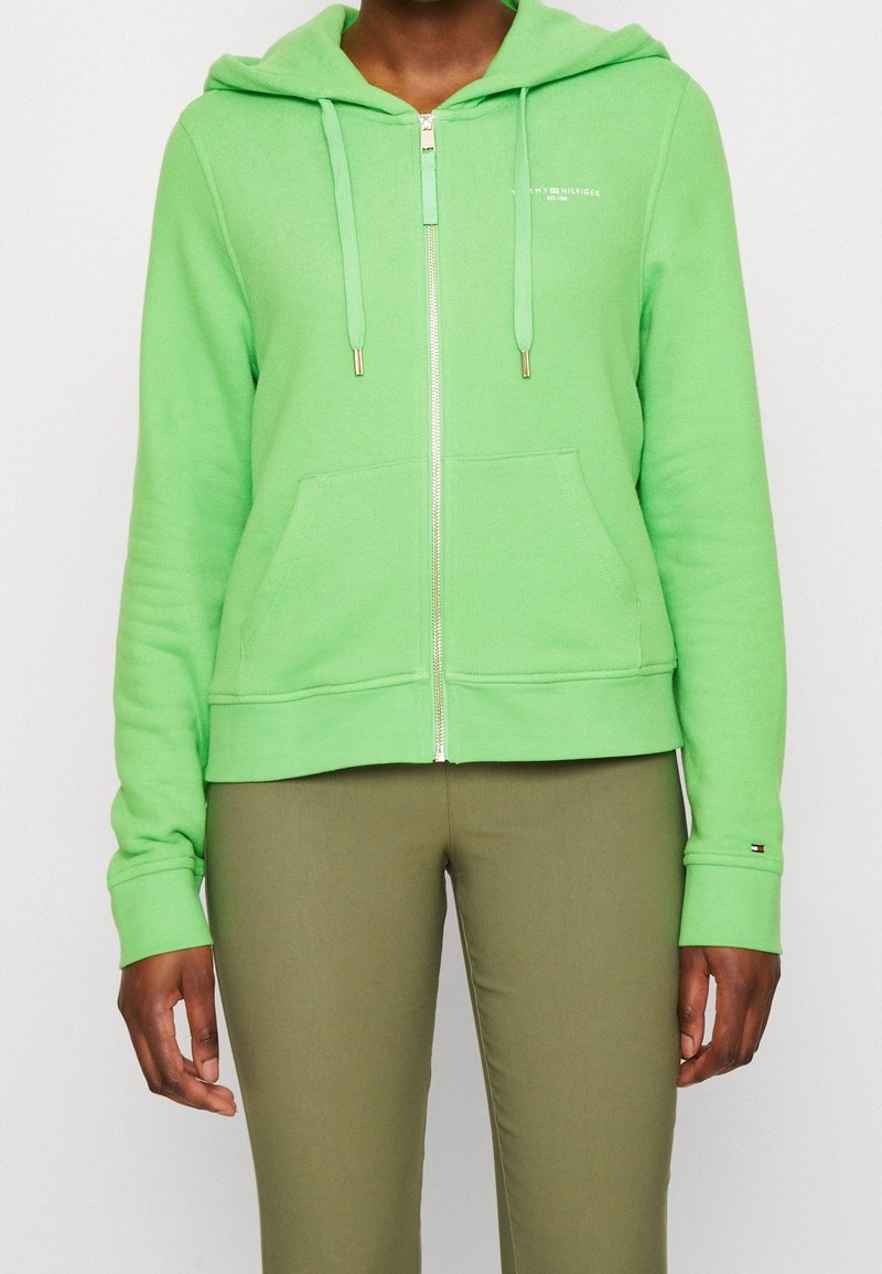 Sudadera verde con cremallera, hecha de tejido suave, que cuenta con un bolsillo frontal, cordones y herrajes en tono dorado. Llevada con pantalones verde oliva.