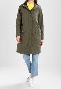 Olivově zelený deštník do pasu s kapucí, s předními patentkami, dvěma velkými náprsními kapsami a volným střihem přes světle modré džíny.