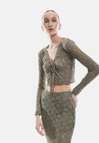 Olijfgroene kanten crop top met lange mouwen, diepe V-hals en sluiting met strik. Gekoppeld aan een aansluitende rok, beide met bloemmotieven.