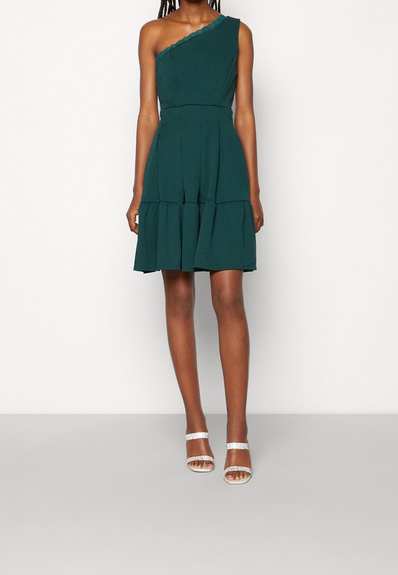 Mannequin portant une robe sans manches vert foncé, asymétrique avec une jupe à volants, associée à des talons blancs à brides.