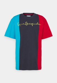 Tričko s barevnými bloky, které obsahuje tyrkysové, námořnické modré a červené sekce. Na přední straně je žlutý kurzivní text. Krátký rukáv, volný střih.