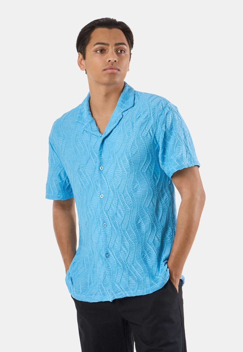 Jeune homme portant une chemise à boutons bleu clair texturée à manches courtes avec col, debout les mains dans les poches d'un pantalon noir.