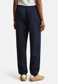Person trägt dunkelblaue Jogginghose, weißes T-Shirt und beige Sneakers, von hinten vor weißem Hintergrund gezeigt.