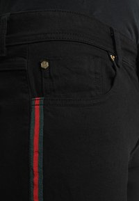 Jeans de mezclilla negra con un diseño ajustado, detalles en hardware dorado y una franja lateral en colores verde y rojo. Textura suave.