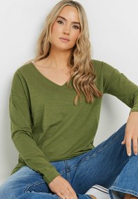Olivegrünes Langarmoberteil mit V-Ausschnitt. Weicher Stoff, lockere Passform, kombiniert mit blauen Jeans und einer sitzenden Pose.