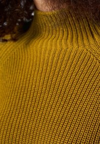 Gros plan sur un pull à col roulé côtelé couleur moutarde porté par une personne aux cheveux bouclés foncés.