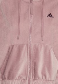 Veste zippée rose en velours côtelé. Dotée d'une capuche, de poches avant et d'un logo Adidas noir brodé sur la poitrine.