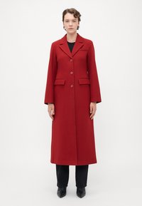 Woman wearing a long red buttoned coat over black pants and black pointed shoes, standing against a plain white background.