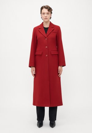 Woman wearing a long red buttoned coat over black pants and black pointed shoes, standing against a plain white background.