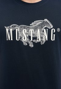 Tumši zils kokvilnas T-krekls ar baltu grafiku, kurā attēlots skrienošs zirgs, un virs tā drukāts vārds "MUSTANG" treknajiem burtiem.