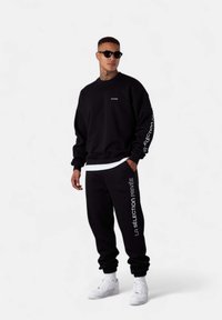 Ensemble en sweat-shirt et pantalon de jogging noir, avec des accents de texte et de logo blancs. Le matériau semble doux ; des lunettes de soleil complètent le look décontracté.