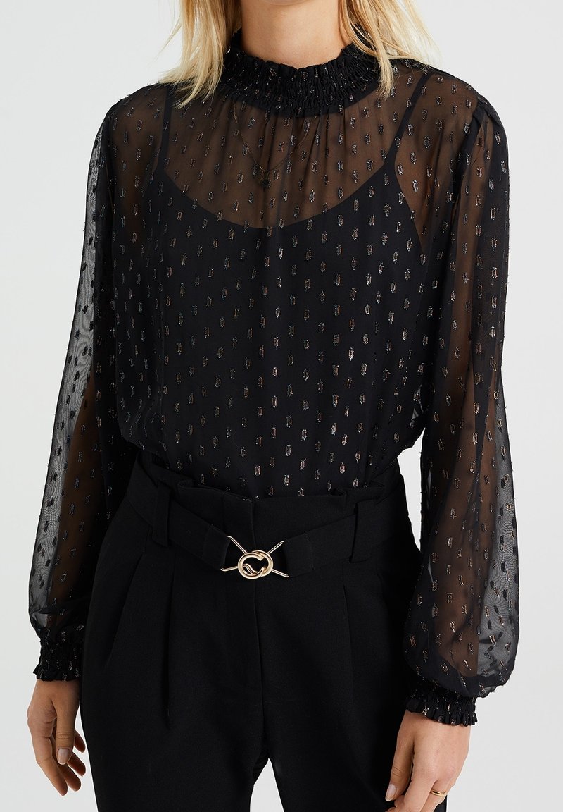 Femme portant un chemisier noir transparent à pois métalliques, col et poignets volantés, rentré dans un pantalon noir taille haute avec une boucle de ceinture dorée.
