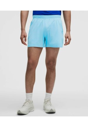 Mann trägt hellblaue Sportsshorts, weiße Socken und weiße Laufschuhe und steht vor einem schlichten Hintergrund.