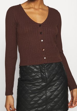 Femme portant un cardigan marron côtelé à boutons et une jupe en cuir matelassé noire, présentée du cou jusqu'à mi-cuisse.