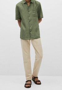 Zielona koszula z krótkim rękawem, jasnobeżowe spodnie i brązowe sandały. Koszula posiada kieszeń, a spodnie mają luźny krój.