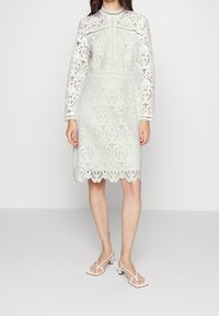 Krajková šaty v jemné mátově zelené barvě, s dlouhými rukávy, vlnkovým lemováním a složitými květinovými vzory. Spárovány s minimalistickými bílými sandály.