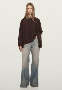 Donna che indossa un poncho largo di colore marrone scuro e jeans a gamba larga di colore blu chiaro, in piedi con una mano nella tasca contro uno sfondo neutro.