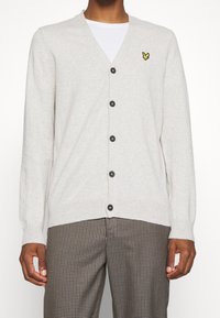 Hellgrauer Cardigan mit V-Ausschnitt, knöpfbarer Front und langen Ärmeln. Besitzt ein kleines gelbes Logo auf der linken Brust und dunkle Knöpfe.