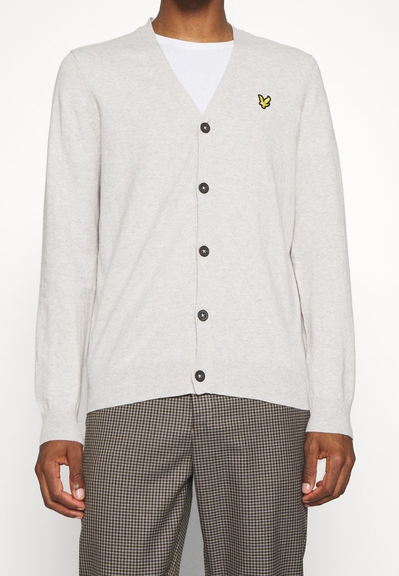 Hellgrauer Cardigan mit V-Ausschnitt, knöpfbarer Front und langen Ärmeln. Besitzt ein kleines gelbes Logo auf der linken Brust und dunkle Knöpfe.