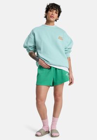 Πράσινη φαρδιά φούτερ με στρογγυλή λαιμόκοψη και χρυσό λογότυπο, σετ με πράσινα σορτς, ροζ κάλτσες και μπεζ σανδάλια slide.