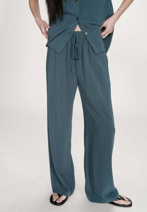 Femme portant un pantalon ample teal avec cordon de serrage et une chemise assortie à boutons, debout les mains près des hanches, portant des tongs noires.