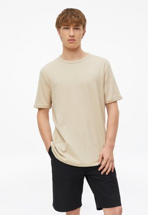 Młody mężczyzna stoi z rękami w kieszeniach, ubrany w beżowy t-shirt z krótkim rękawem i czarne szorty na jednolitym białym tle.
