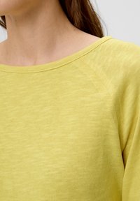 Gelbes Strickoberteil mit weitem Rundhalsausschnitt und Raglanärmeln. Weiche Textur mit einem dezenten melierten Muster.