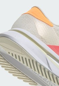 Light beige running shoe with orange and coral accents, textured mesh upper, white midsole, and a flexible rubber outsole with grooves.