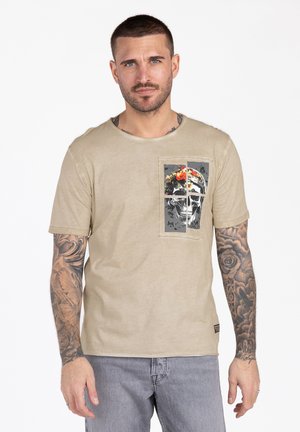 Uomo con capelli corti e scuri e tatuaggi su entrambe le braccia, che indossa una t-shirt beige con grafica di teschi e fiori e jeans grigio chiaro, in piedi con espressione neutra.