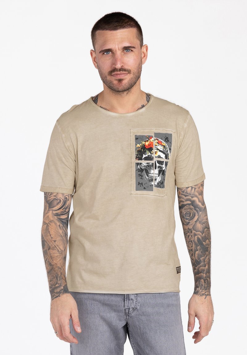Mann mit kurzen dunklen Haaren und Tätowierungen auf beiden Armen, der ein beiges T-Shirt mit Schädel- und Blumenmotiv und hellgraue Jeans trägt, steht neutral.