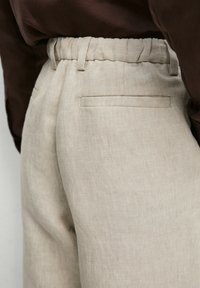 Close-up of beige linen pants with elastic waistband and back slit pocket, worn with a dark brown top.