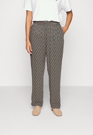 Osoba nosząca luźne spodnie w geometryczne wzory w kolorze czarnym i beżowym z elastycznym pasem, biały krótki rękaw oraz beżowe plecione wsuwane buty.