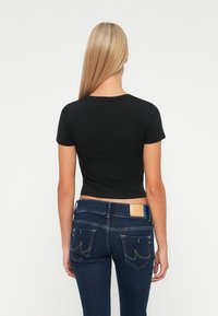 Mujer con cabello rubio largo, vistiendo una blusa negra ajustada de manga corta y jeans de mezclilla azul oscuro, de espaldas contra un fondo sencillo.