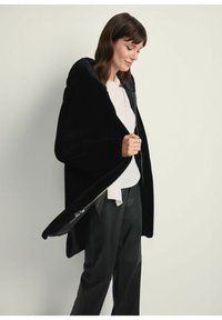 Czarny, pluszowy płaszcz z kapturem, zapinany na zamek, z szerokimi rękawami i gładką fakturą. Noszony swobodnie na białej bluzce i czarnych spodniach.