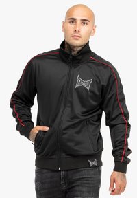 Chaqueta negra con cremallera y rayas laterales rojas, con el logo de TAPOUT. Presenta un cuello, dos bolsillos laterales y puños acanalados. Hecha de un tejido brillante.